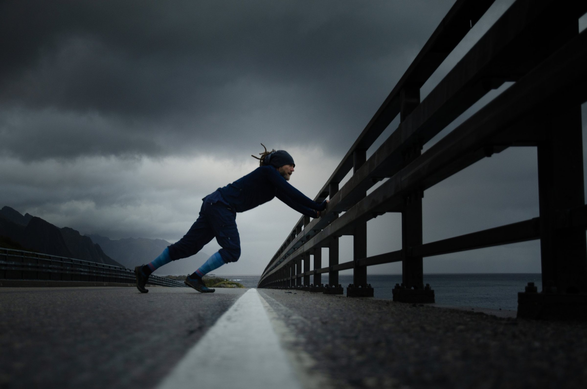 Marc s’étire contre la rambarde d’un pont, bas de compression bleus aux jambes, sous un ciel d’orage, face aux montagnes et à la mer.