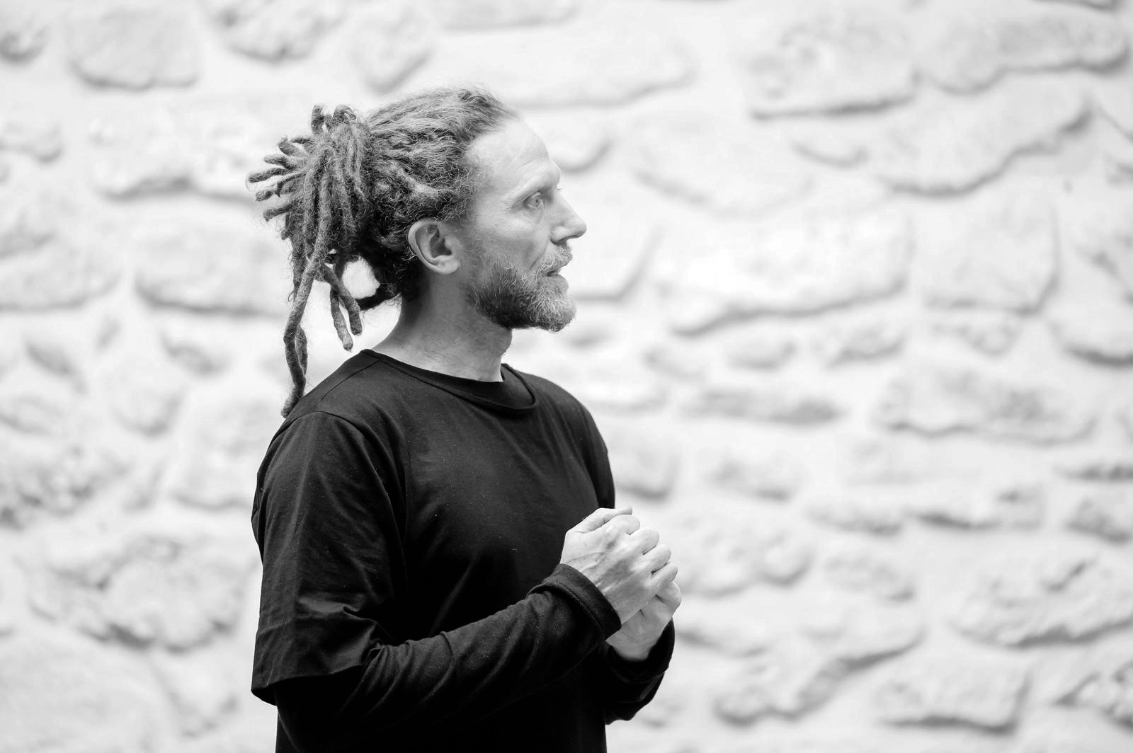 Portrait de Marc Papillon de profil, mains jointes, devant un mur de pierres blanches.